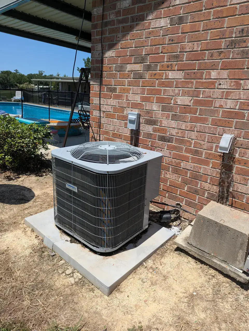 Outdoor air conditioning unit on a square pad beside a brick wall and patio